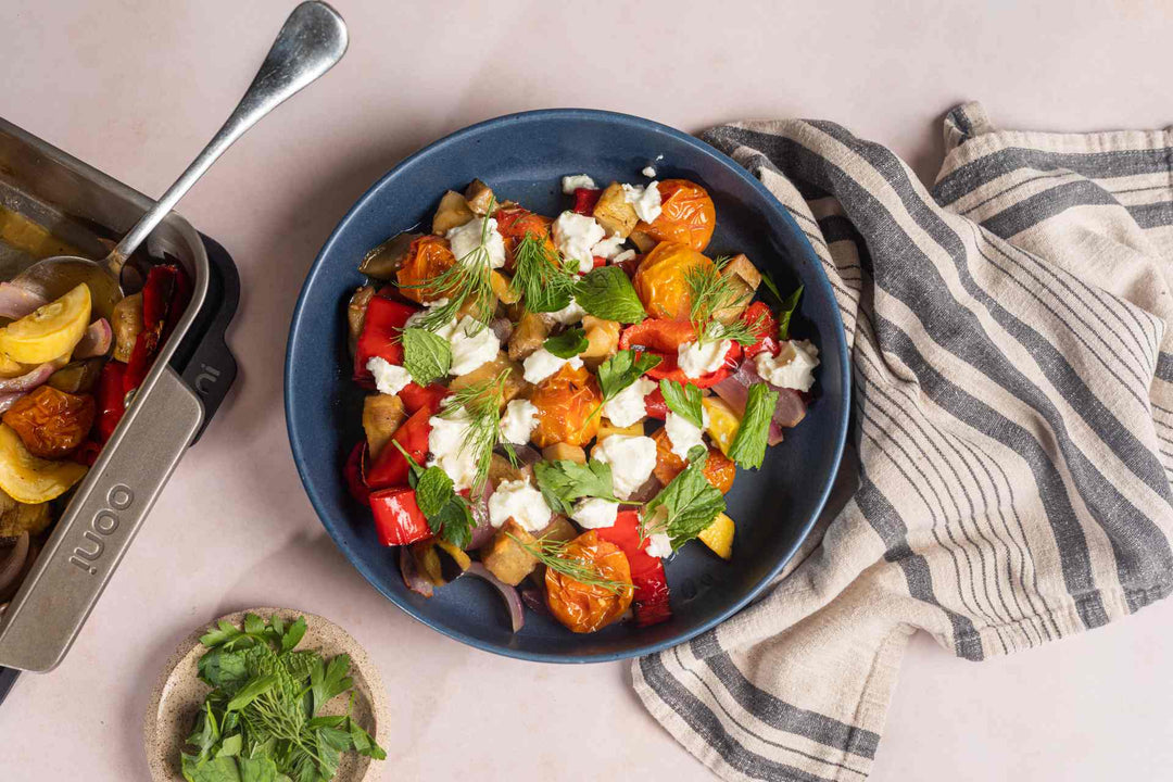 Slow-roasted squash, eggplant, onions, cherry tomatoes and sweet capsicums in an Ooni Roasting pan next to a bowl of slow-roasted vegetables with feta and fresh herbs on a table.