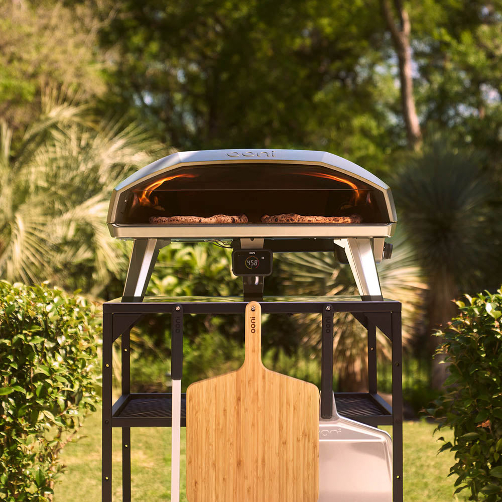 An outdoor setup featuring an Ooni Koda 2 Max pizza oven on a black stand. The oven top shows the 'ooni' logo, and a digital display reads '458'. A wooden pizza peel with the 'ooni' logo is positioned in the foreground.
