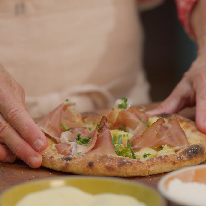  Two hands touching a cooked pizza with speck, pineapple and chives.