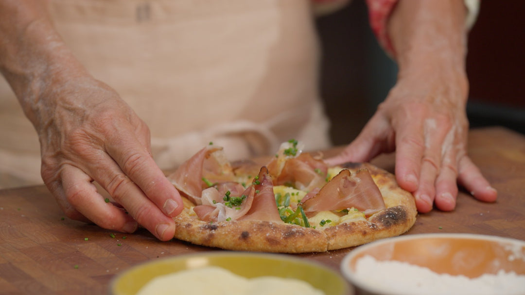  Two hands touching a cooked pizza with speck, pineapple and chives.