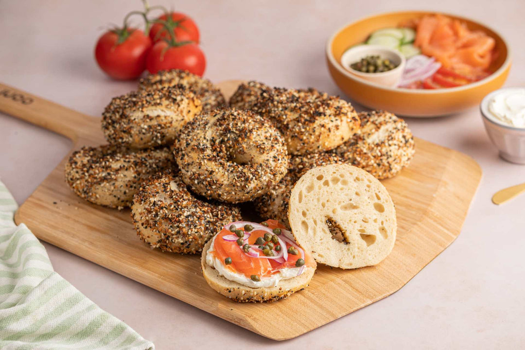 A stack of unsliced everything bagels and one bagel sliced and spread with cream cheese, smoked salmon, onions and capers on an Ooni Bamboo Pizza Peel 