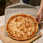 signature perforated pizza peel placing pizza on bamboo pizza peel and serving board.